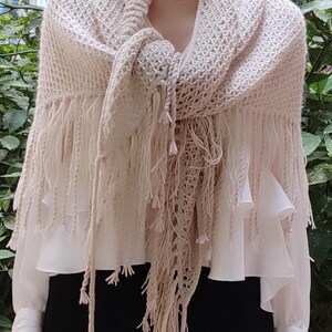 May include: A beige knit shawl featuring a textured, open-weave pattern and long, dangling fringe along the edges. The shawl is draped over a white long-sleeved blouse with ruffled details and a solid black garment worn underneath.