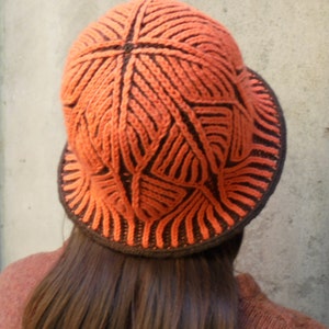 Puede incluir: Un gorro de punto con un patrón geométrico marrón y naranja. El gorro está hecho de un hilo suave y texturizado y tiene un ala ancha.