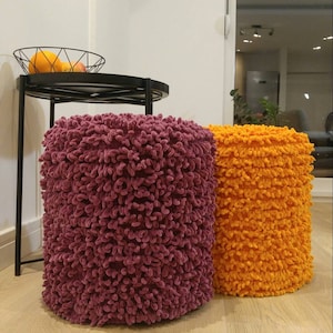 Könnte beinhalten: Zwei runde, flauschige Hocker, einer in einem tiefen Lila und der andere in einem leuchtenden Orange. Die Hocker sind aus einem weichen, strukturierten Stoff gefertigt und eignen sich perfekt, um jedem Raum einen Hauch von Farbe und Komfort zu verleihen.