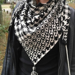 Puede incluir: Un chal triangular blanco y negro con un patrón geométrico y una borla negra. El chal lo lleva una persona que lleva una camisa negra y una chaqueta de cuero negra.