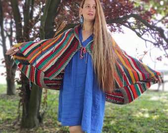 May include: A woman wearing a colorful striped crocheted shawl with a blue dress. The shawl has a fringe trim and is made with a variety of colors, including red, orange, yellow, green, blue, and purple.
