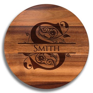 May include: A round wooden cutting board with a dark brown finish. The board features an engraved letter "S" with floral detailing above and below the word "SMITH". The design is centered on the board.