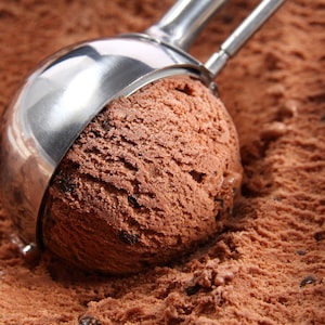 May include: A close-up of a silver ice cream scoop scooping out a large portion of chocolate ice cream with chocolate chips. The ice cream is in a container and the scoop is partially submerged in the ice cream.