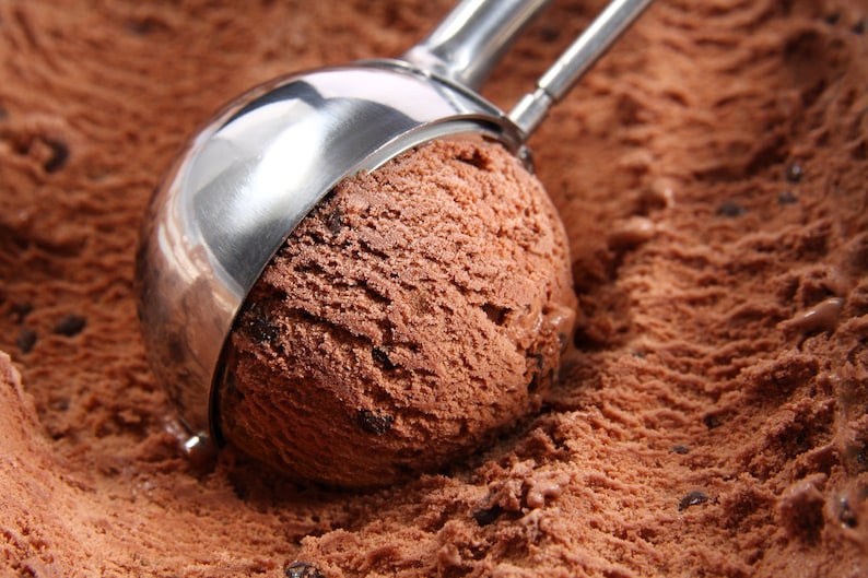 May include: A close-up of a silver ice cream scoop scooping out a large portion of chocolate ice cream with chocolate chips. The ice cream is in a container and the scoop is partially submerged in the ice cream.