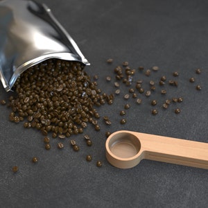 May include: A silver foil bag spills out dark brown coffee beans onto a dark gray surface. A wooden scoop with a clear plastic insert sits on the surface.