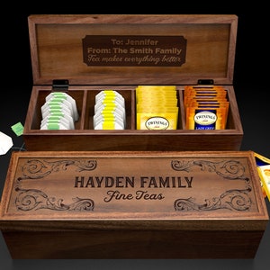 May include: A wooden tea box with a personalized inscription that reads "Hayden Family Fine Teas". The box has four compartments for tea bags and a lid that lifts to reveal the tea bags. The lid has a personalized inscription that reads "To: Jennifer From: The Smith Family Tea makes everything better".