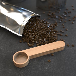 May include: A wooden coffee scoop with a rounded handle and a shallow bowl. The scoop is sitting on a dark gray surface with coffee beans scattered around it.
