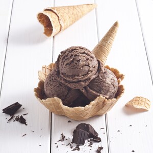 May include: Three scoops of chocolate ice cream in a waffle cone bowl with a waffle cone on the side and chocolate shavings on a white wooden surface.