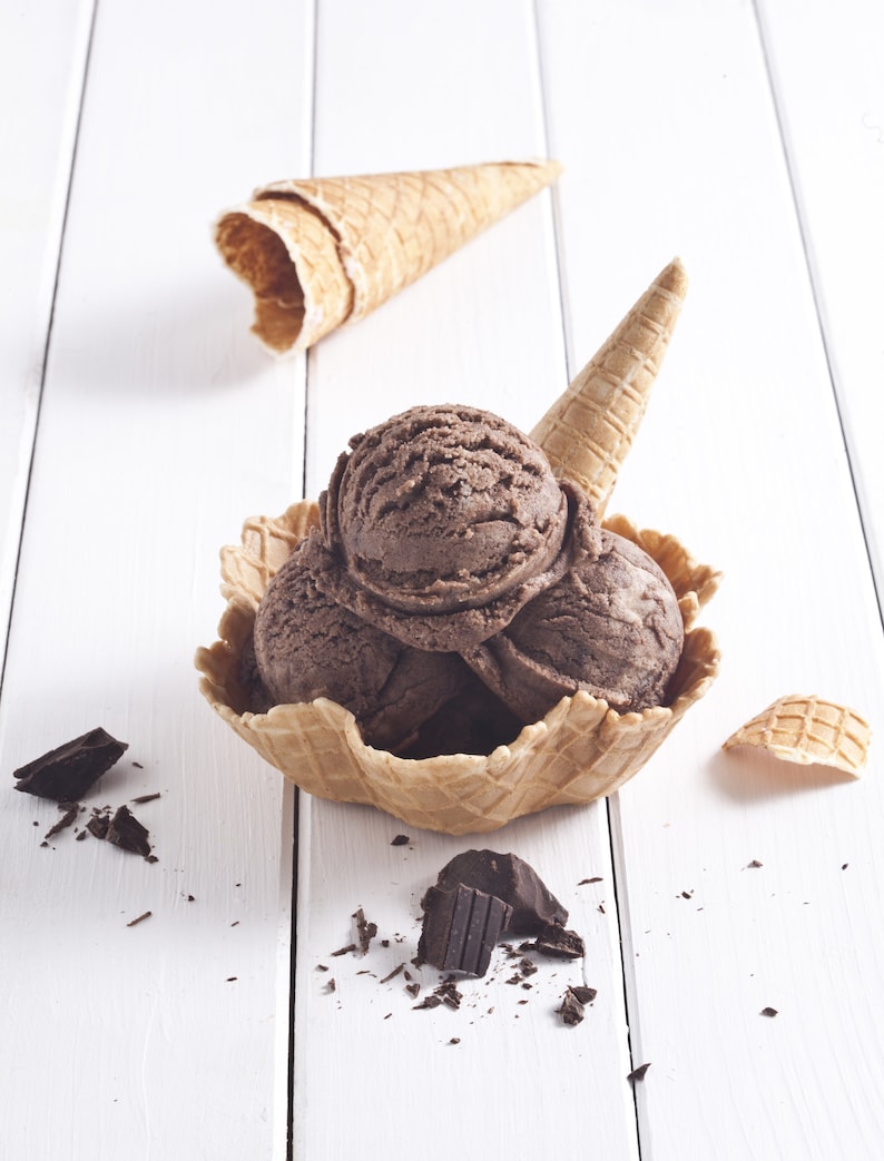 May include: Three scoops of chocolate ice cream in a waffle cone bowl with a waffle cone on the side and chocolate shavings on a white wooden surface.