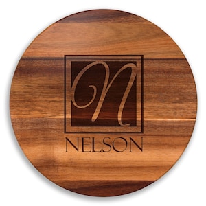 May include: A round wooden cutting board with a dark brown finish. The center features a square with the letter "N" in an elegant script, and the name "NELSON" below it, all in a darker tone.