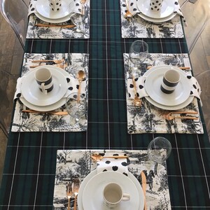 May include: A green and black plaid tablecloth with six place settings. Each place setting includes a white plate, a white cup, a black and white napkin, a gold fork, a gold knife, and a gold spoon. The tablecloth has a black and white pattern with a horse and carriage design.