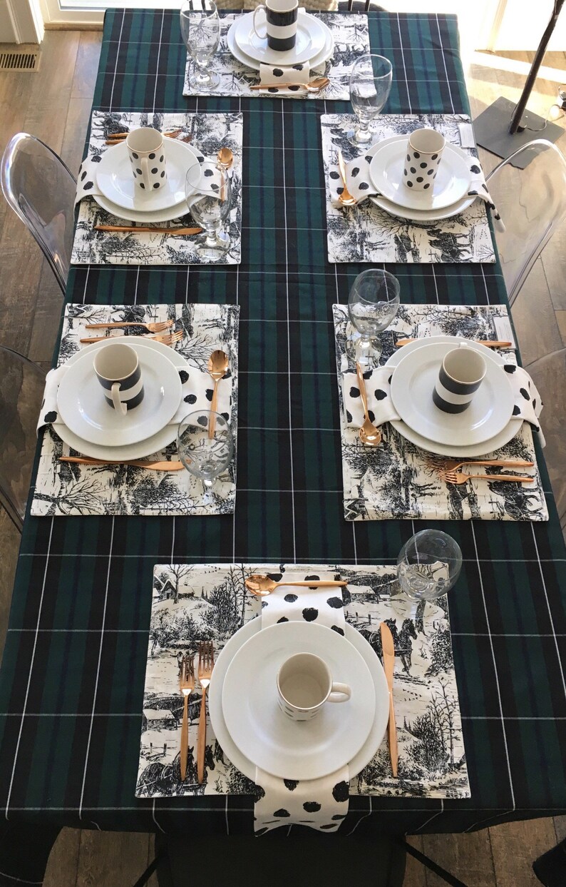 May include: A green and black plaid tablecloth with six place settings. Each place setting includes a white plate, a white cup, a black and white napkin, a gold fork, a gold knife, and a gold spoon. The tablecloth has a black and white pattern with a horse and carriage design.