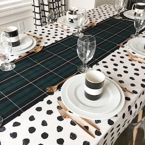 Pode incluir: Um corredor de mesa xadrez preto e branco com uma toalha de mesa de bolinhas pretas e brancas por baixo. A mesa está posta com pratos brancos, talheres dourados e canecas pretas e brancas.