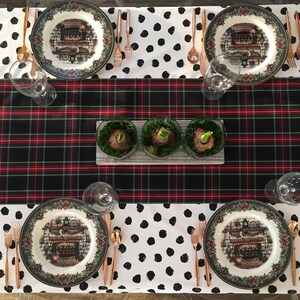 Pode incluir: Uma toalha de mesa xadrez preta e vermelha com uma toalha de mesa branca com bolinhas pretas por cima. Existem quatro lugares à mesa com pratos que representam uma cena de inverno e uma lareira. Há três pequenos recipientes de vidro com plantas verdes no centro da mesa.
