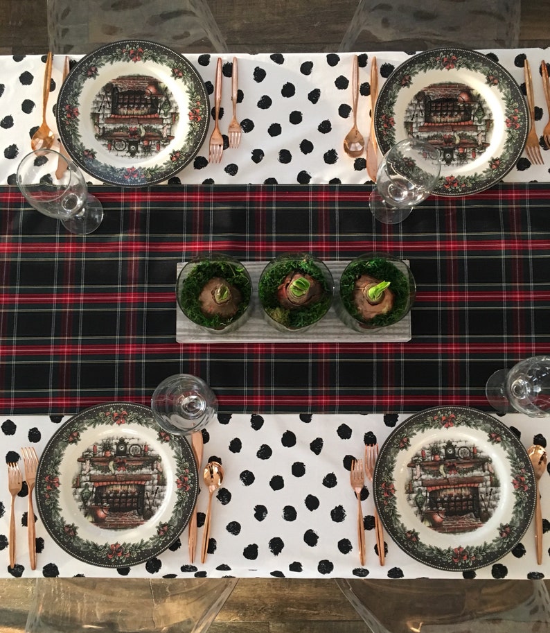 May include: A black and red plaid tablecloth with a white tablecloth with black polka dots on top. There are four place settings with plates featuring a winter scene and a fireplace. There are three small glass containers with green plants in the center of the table.