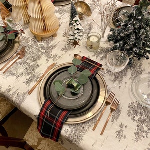 May include: A white tablecloth with a black and white pattern of trees and flowers. The table is set with black and silver plates, cutlery, and glasses. There are also small Christmas trees and other decorations on the table.