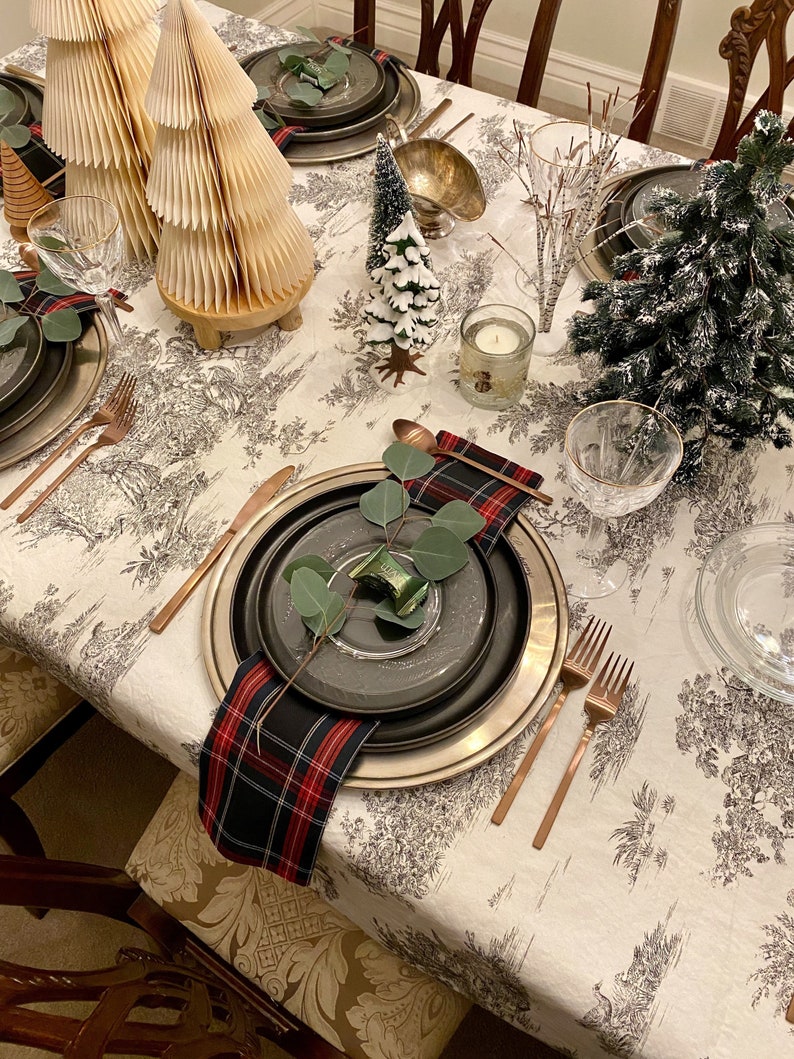 May include: A white tablecloth with a black and white pattern of trees and flowers. The table is set with black and silver plates, cutlery, and glasses. There are also small Christmas trees and other decorations on the table.
