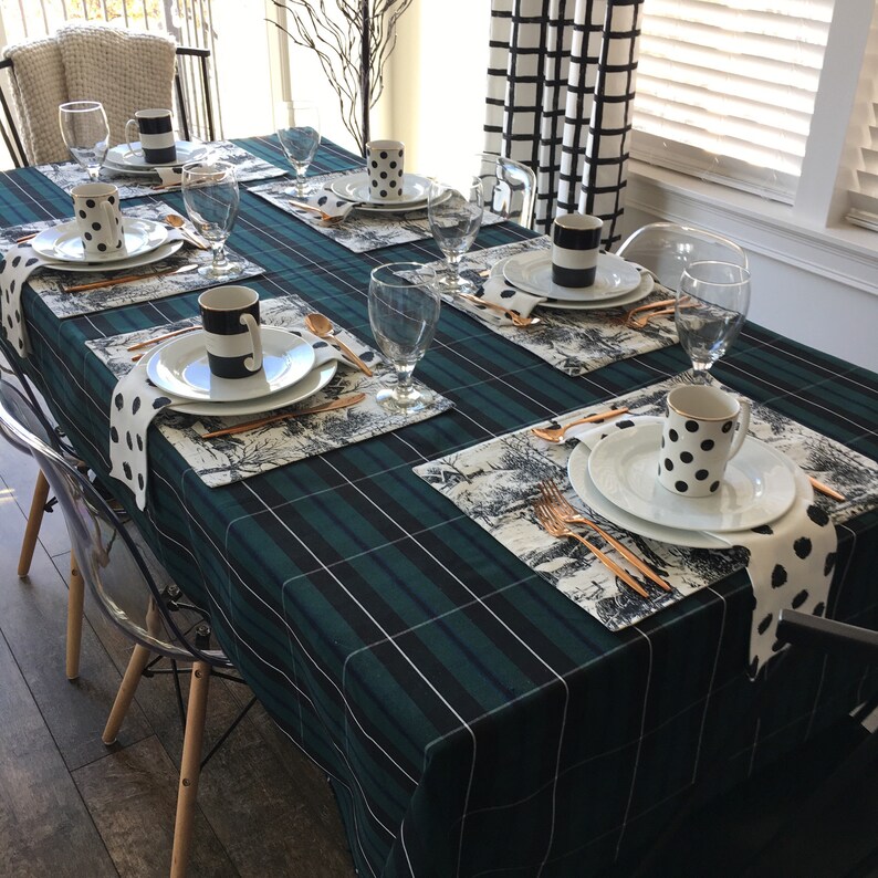 May include: A green and white plaid tablecloth with black and white patterned placemats and napkins. Each place setting includes a white plate, a black and white mug, a gold fork and knife, and a clear glass.