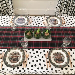 May include: A black and white polka dot tablecloth with a red and black plaid runner. There are three clear glass vases with green moss and white bulbs on the runner. There are four place settings with white plates with a green and red border and a Christmas scene in the center. Each place setting has a glass wine glass, a fork, a knife, and a spoon.