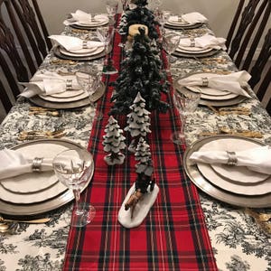 May include: A red and black plaid table runner with a white background. The runner is decorated with small, snow-covered pine trees and a miniature horse-drawn carriage. The table is set with white plates, silver chargers, and white napkins.