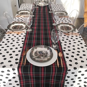 May include: A black and red plaid table runner with a white background with black polka dots. The table runner is set on a table with a white tablecloth. There are six place settings on the table, each with a white plate, a green and brown patterned bowl, a glass, and gold silverware.