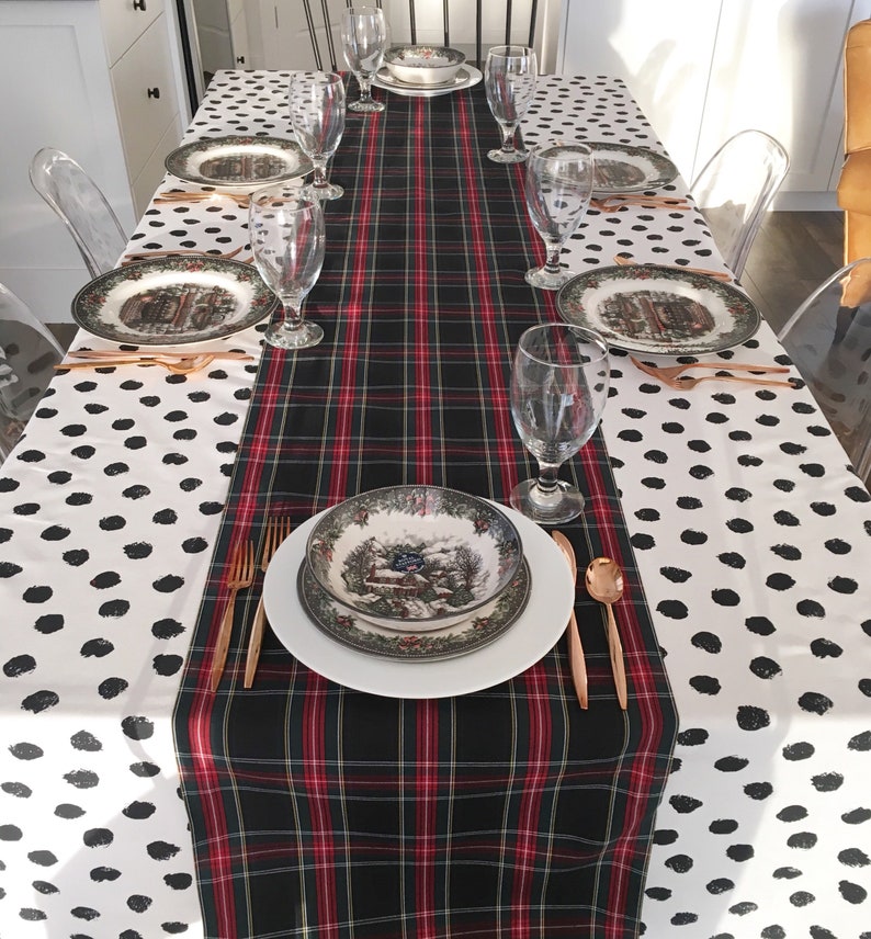 May include: A black and red plaid table runner with a white background with black polka dots. The table runner is set on a table with a white tablecloth. There are six place settings on the table, each with a white plate, a green and brown patterned bowl, a glass, and gold silverware.