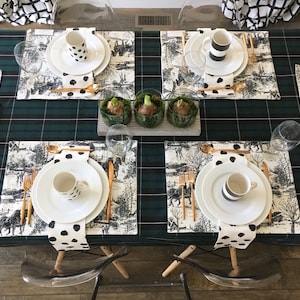 May include: A green and black plaid tablecloth with four place settings. Each place setting includes a white plate, a white mug with black polka dots, a white napkin with black polka dots, gold flatware, and a glass. There are three small glass vases with green plants in the center of the table.