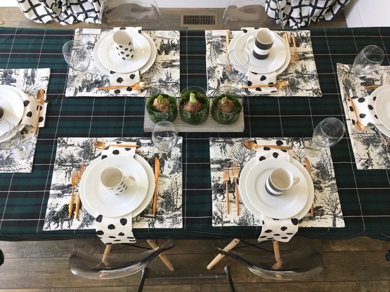 May include: A green and black plaid tablecloth with four place settings. Each place setting includes a white plate, a white mug with black polka dots, a white napkin with black polka dots, gold flatware, and a glass. There are three small glass vases with green plants in the center of the table.