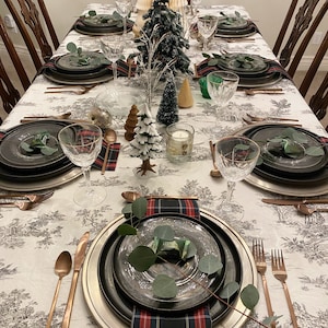 May include: A Christmas table setting with a white tablecloth featuring a black and white pattern. The table is set with black and silver chargers, black plates, and clear glass wine glasses. There are also sprigs of greenery and plaid napkins on the table.