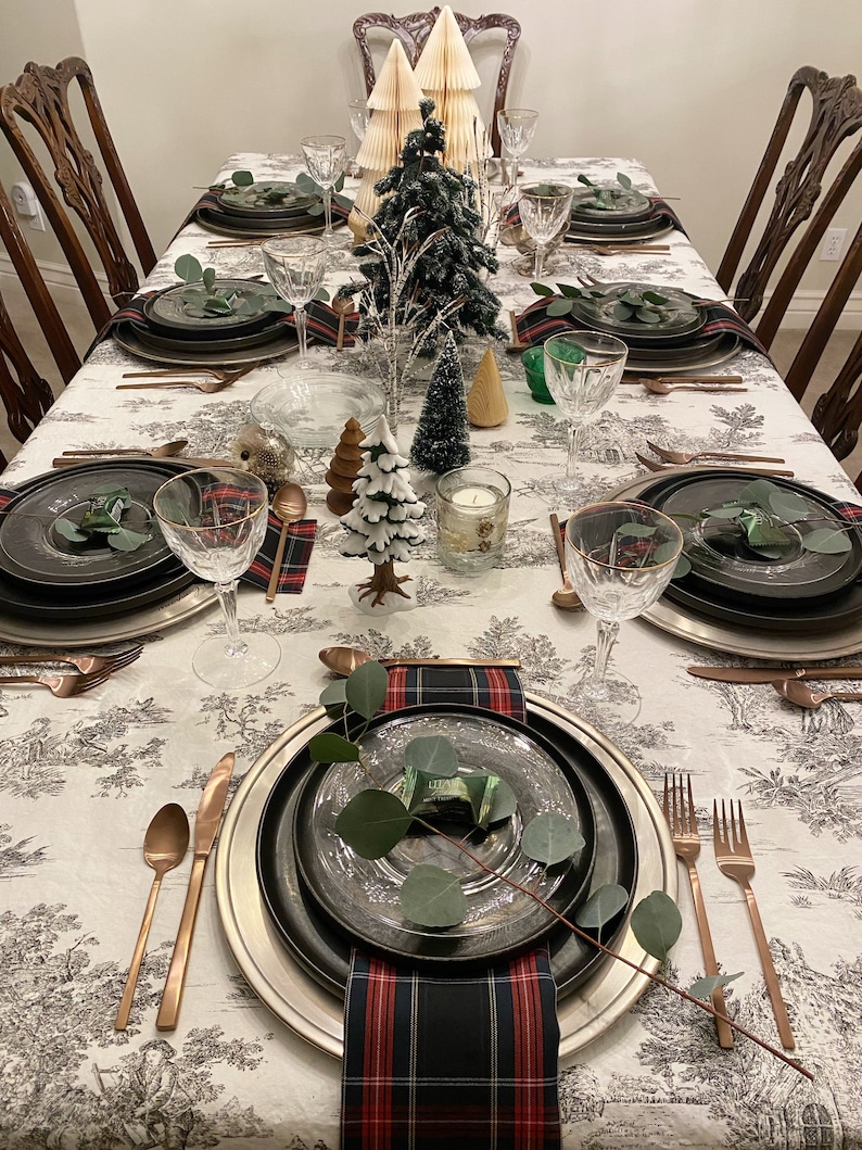 May include: A Christmas table setting with a white tablecloth featuring a black and white pattern. The table is set with black and silver chargers, black plates, and clear glass wine glasses. There are also sprigs of greenery and plaid napkins on the table.