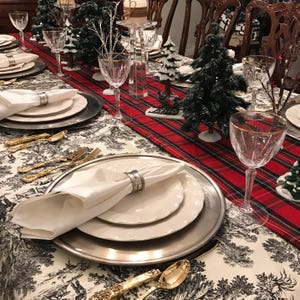 May include: A white napkin with a silver napkin ring is placed on a silver charger plate with a white dinner plate on top. The plates are set on a table with a red and black plaid tablecloth. There are two small artificial Christmas trees on the table, and a glass of water is in the background.