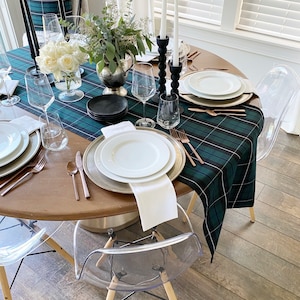 May include: A wooden dining table set with a green and black plaid tablecloth. The table is set with white plates, gold silverware, and clear glasses. There are two black candle holders with white candles on the table. The table is surrounded by four clear acrylic chairs.