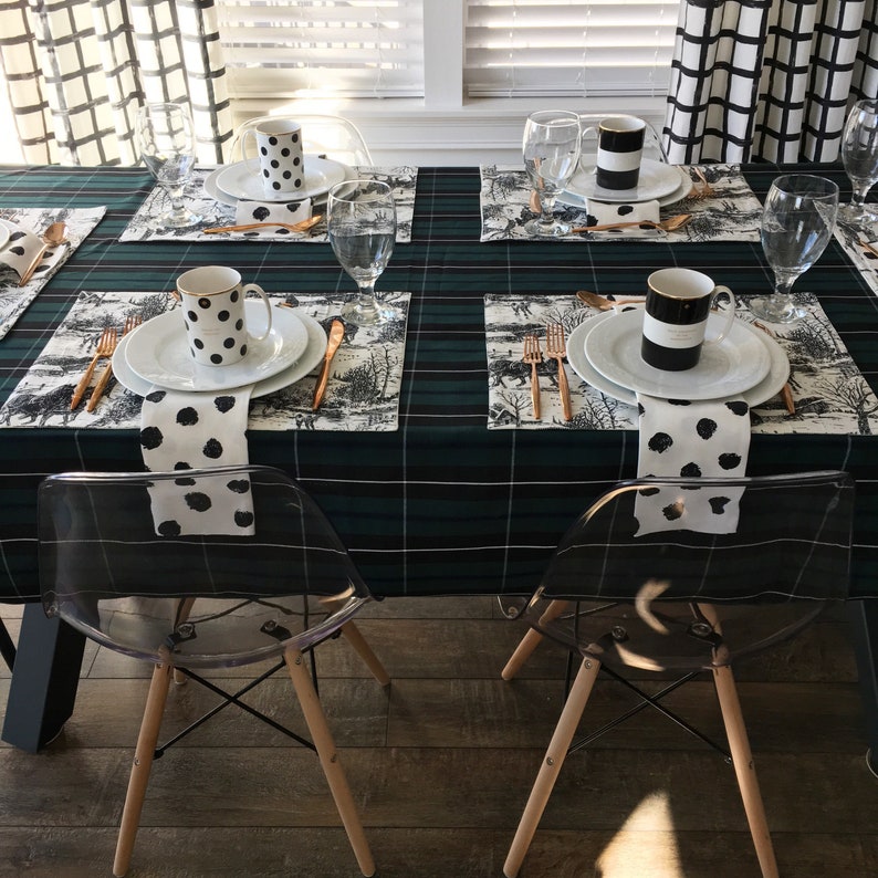 May include: A table setting with a green and black plaid tablecloth. There are four place settings with white plates, black and white napkins, and black and white mugs. The table is set with gold flatware and black and white patterned placemats.