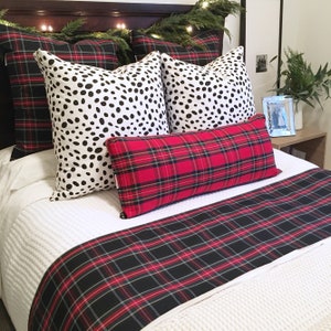 May include: A white bed with a red and black plaid duvet cover and matching pillow. Two black and white spotted pillows are on top of the duvet. The bed is decorated with a garland of greenery and lights.