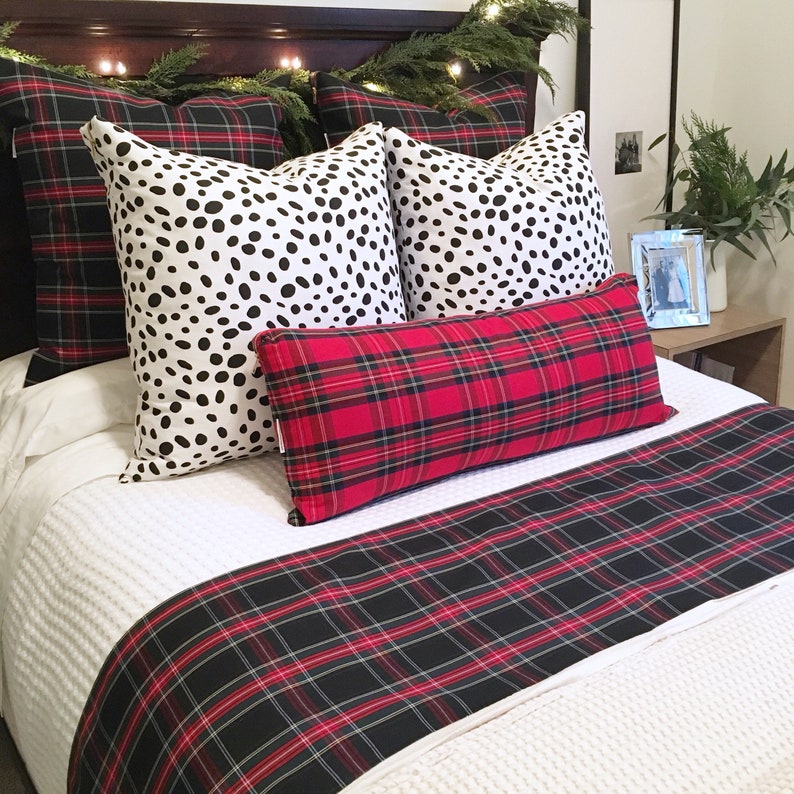 May include: A white bed with a red and black plaid duvet cover and matching pillow. Two black and white spotted pillows are on top of the duvet. The bed is decorated with a garland of greenery and lights.