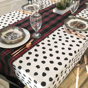 May include: A white tablecloth with black polka dots and a black and red plaid table runner. The table is set with two place settings, each with a white plate, a bowl with a holiday design, and gold silverware.
