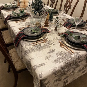 May include: A white tablecloth with a black and white toile pattern is set for a festive dinner. The table is decorated with small artificial pine trees, a paper Christmas tree, and red and black checked napkins. There are also several wine glasses and cutlery on the table.
