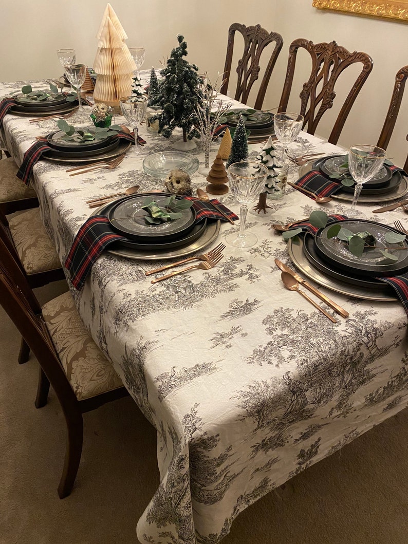 May include: A white tablecloth with a black and white toile pattern is set for a festive dinner. The table is decorated with small artificial pine trees, a paper Christmas tree, and red and black checked napkins. There are also several wine glasses and cutlery on the table.