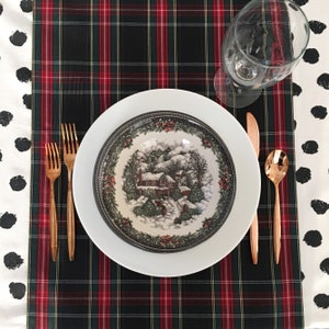 May include: A white dinner plate with a black rim and a winter scene design. The plate is set on a black and red plaid tablecloth with gold flatware and a wine glass.