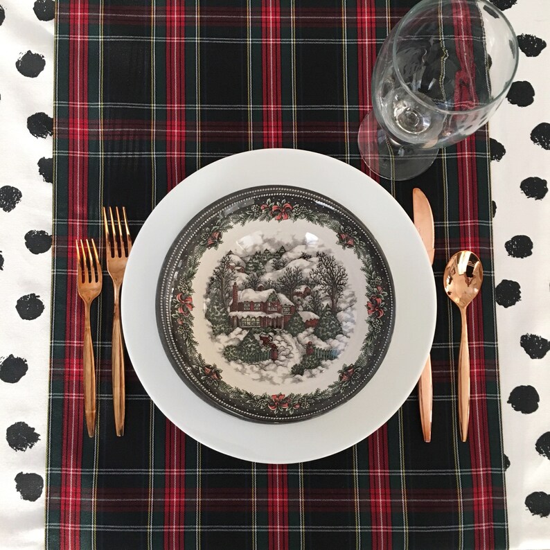 May include: A white dinner plate with a black rim and a winter scene design. The plate is set on a black and red plaid tablecloth with gold flatware and a wine glass.