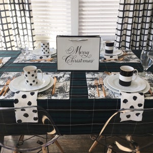 May include: A table setting with a black and white plaid tablecloth, black and white napkins, and black and white polka dot mugs. The table is set for four people with placemats, silverware, and glasses. A white gift bag with the words "Merry Christmas" is on the table.