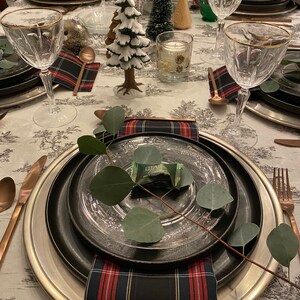 May include: A table setting with a black and silver charger plate, a clear glass plate, a plaid napkin, and a sprig of greenery with a small wrapped treat. The table is decorated with a white and green Christmas tree, a small wooden tree, and a candle.