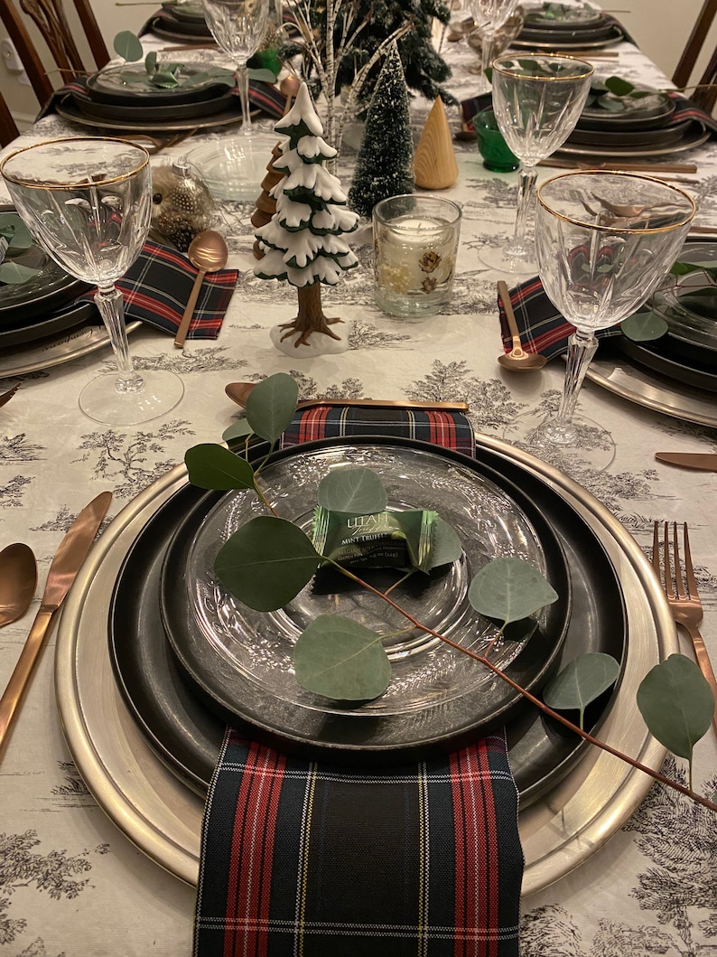 May include: A table setting with a black and silver charger plate, a clear glass plate, a plaid napkin, and a sprig of greenery with a small wrapped treat. The table is decorated with a white and green Christmas tree, a small wooden tree, and a candle.