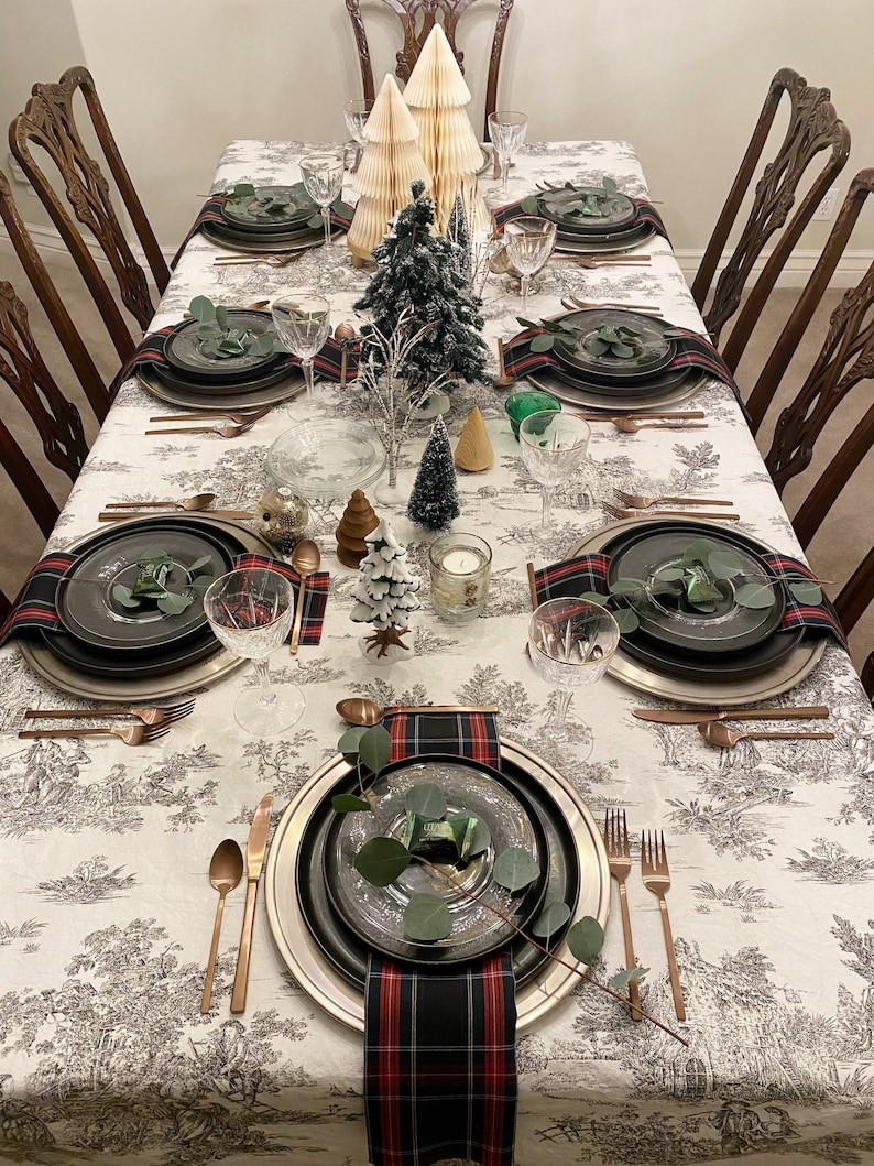 May include: A festive table setting with a white tablecloth featuring a black and white pattern. The table is laid with black and silver plates, copper cutlery, and tartan napkins. Small artificial Christmas trees and greenery are used as centrepieces.