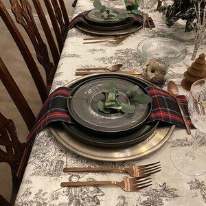 May include: A table setting with a white tablecloth featuring a black and white pattern. The table is set with black and silver plates, copper cutlery, and a plaid napkin. There is a small green plant on each plate.