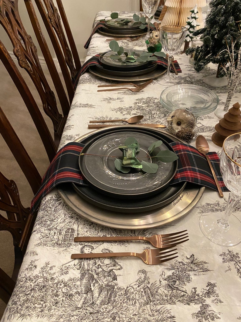May include: A table setting with a white tablecloth featuring a black and white pattern. The table is set with black and silver plates, copper cutlery, and a plaid napkin. There is a small green plant on each plate.