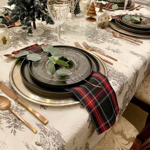 May include: A festive table setting featuring a black and red checked napkin, a silver charger plate, a black plate, and a clear glass plate. A sprig of greenery rests on the plate. The table is adorned with a white tablecloth with a black and white pattern.