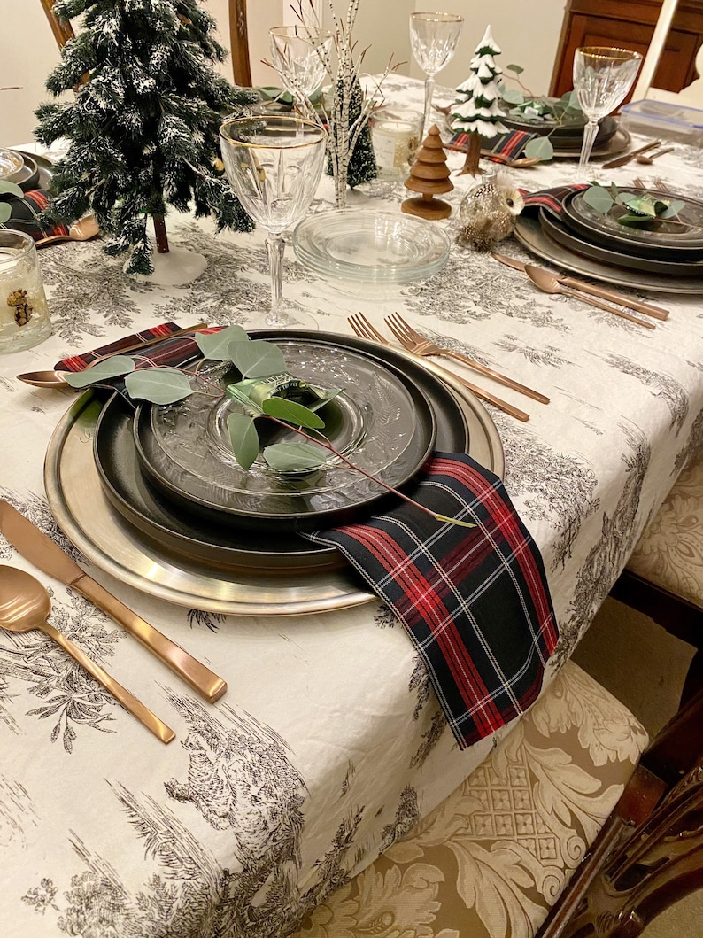 May include: A festive table setting featuring a black and red checked napkin, a silver charger plate, a black plate, and a clear glass plate. A sprig of greenery rests on the plate. The table is adorned with a white tablecloth with a black and white pattern.
