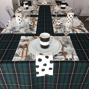 Pode incluir: Uma toalha de mesa xadrez verde e preta com placemats brancos e guardanapos de bolinhas pretas e brancas. Há canecas de bolinhas pretas e brancas, pratos brancos e talheres dourados na mesa.