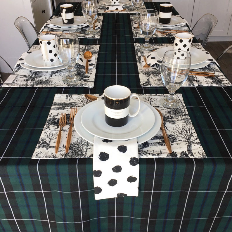 May include: A green and black plaid tablecloth with white placemats and black and white polka dot napkins. There are black and white polka dot mugs, white plates, and gold silverware on the table.